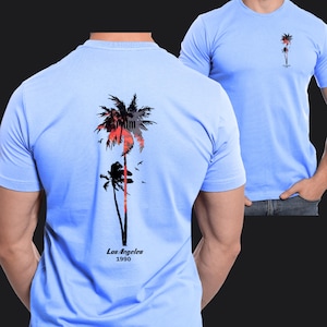 Pu&ograve; includere: T-shirt azzurro con una grafica di palma sul retro e una versione pi&ugrave; piccola sul davanti. La grafica include palme rosse e nere, uccelli e la scritta "Los Angeles 1990".