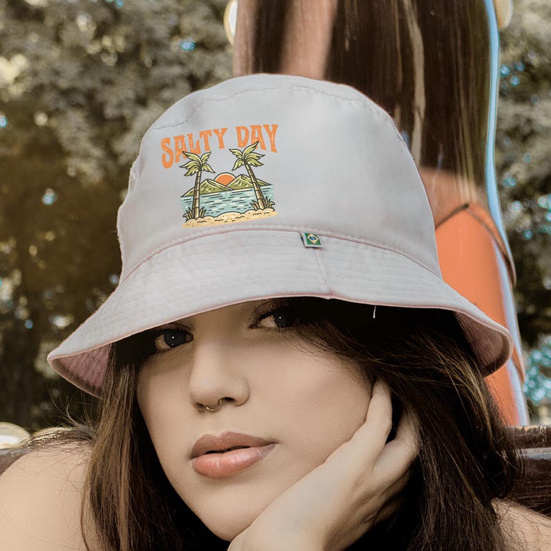 Pu&ograve; includere: Cappello a secchiello rosa chiaro con la scritta "Salty Day" in arancione, raffigurante una scena di spiaggia con palme e un tramonto. Il cappello ha un piccolo dettaglio quadrato verde sul lato.