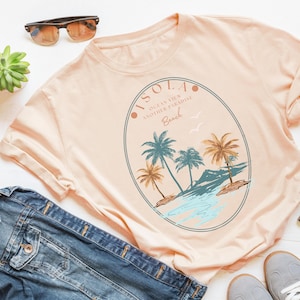 Pu&ograve; includere: Una maglietta color pesca con una grafica di una scena di spiaggia tropicale. La grafica presenta palme, oceano e il testo "ISOLA OCEAN VIEW ANOTHER PARADISE Beach". Occhiali da sole, una giacca di jeans e scarpe da ginnastica sono visibili.