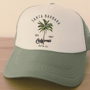 Pu&ograve; includere: Cappellino da camionista bianco e verde con la scritta "Santa Barbara" e una grafica di palma. Il cappellino presenta anche la scritta "California" e "Surf the Sun". Il cappellino ha una visiera curva e un retro in rete.
