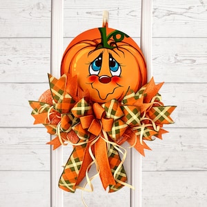 Op de afbeelding: Een decoratieve deurhanger met een ronde oranje pompoenuitsparing met een cartoongezicht, grote blauwe ogen en een groene steel. De pompoen is versierd met een grote, meerlaagse strik gemaakt van oranje en groen geruite stoffen linten.