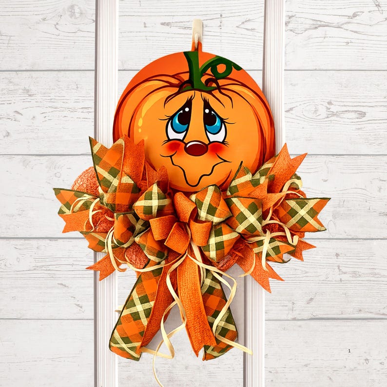 Op de afbeelding: Een decoratieve deurhanger met een ronde oranje pompoenuitsparing met een cartoongezicht, grote blauwe ogen en een groene steel. De pompoen is versierd met een grote, meerlaagse strik gemaakt van oranje en groen geruite stoffen linten.