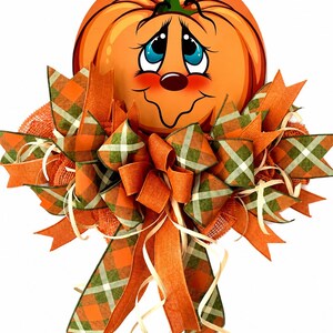 Op de afbeelding: Een decoratieve wandhanger met een cartoonpompoen met een lachend gezicht, blauwe ogen en roze wangen. De pompoen is versierd met een grote, meerlaagse strik gemaakt van oranje jute en groen-oranje geruite stoffen linten met dun raffia.