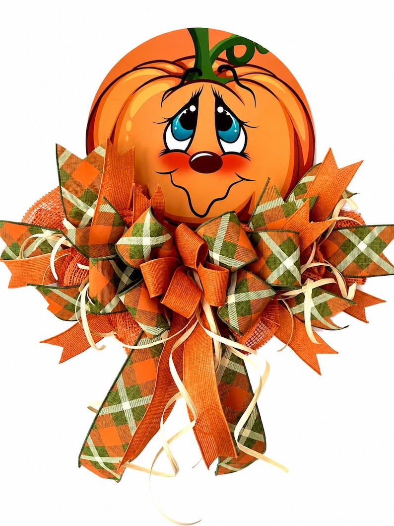 Op de afbeelding: Een decoratieve wandhanger met een cartoonpompoen met een lachend gezicht, blauwe ogen en roze wangen. De pompoen is versierd met een grote, meerlaagse strik gemaakt van oranje jute en groen-oranje geruite stoffen linten met dun raffia.