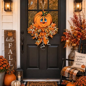 Op de afbeelding: Een decoratieve deurkrans met een geschilderde oranje pompoen met een lachend gezicht, omringd door oranje en groene geruite linten. De krans hangt aan een zwarte deur, vergezeld van een deurmat, pompoenen en een bord met de tekst hello FALL.