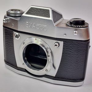 May include: A silver and black Exa 500 35mm film camera body featuring a textured black leatherette grip, a central lens mount, a top-mounted film advance lever, and a shutter release button. The camera has a metallic silver finish.