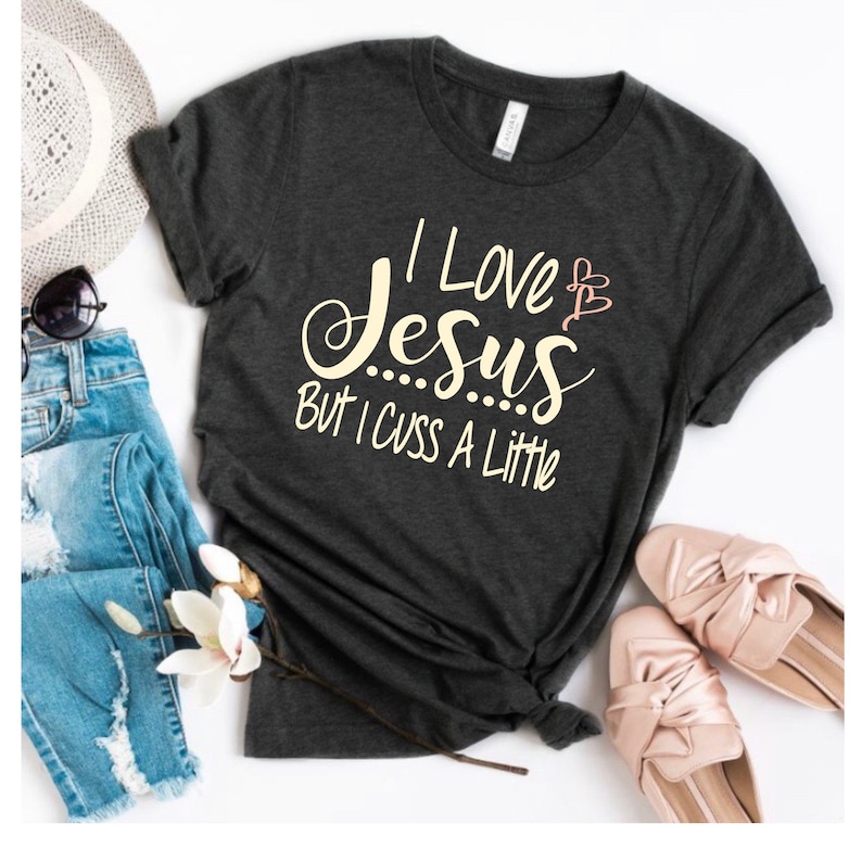May include: A dark gray t-shirt with the text "I Love Jesus But I Cuss A Little" in white lettering. The text is in a handwritten font. There are two pink hearts above the word "Jesus".