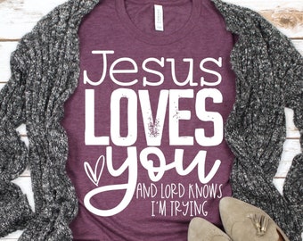 May include: A maroon t-shirt with the text "Jesus Loves You" and "And Lord Knows I'm Trying" in white. The shirt is styled with a gray cardigan, blue jeans, and brown tassel loafers.