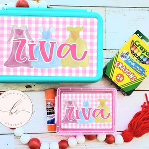May include: A turquoise lunchbox with a pink and white gingham pattern featuring three princess dresses and the name "Ziva". A pink and white gingham pencil box with the name "Ziva" is also shown.