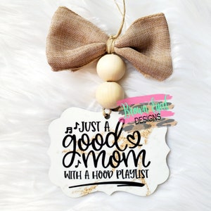 May include: White wooden sign with gold glitter accents and black text that reads "Just a good mom with a hood playlist". The sign is hanging from a brown burlap bow with wooden beads.
