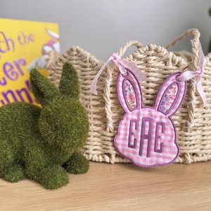 Op de afbeelding: Een groene, met mos bedekte konijnenfiguur staat naast een gevlochten mand met een roze geruite konijnvormige tag. De tag heeft oren met bloemenprint en de letters "GAE". Roze linten zijn aan de mand vastgemaakt.