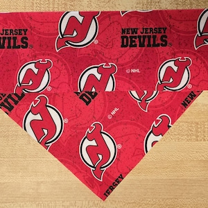 May include: Red fabric with a repeated pattern of the New Jersey Devils logo. The text "NEW JERSEY DEVILS" is printed on the fabric. The fabric is folded in a triangular shape.