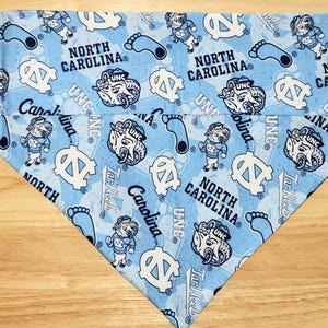 May include: Light blue bandana with a repeating pattern of the words "North Carolina", the letters "UNC", cartoon ram mascots, and footprints. The bandana is folded into a triangle shape.