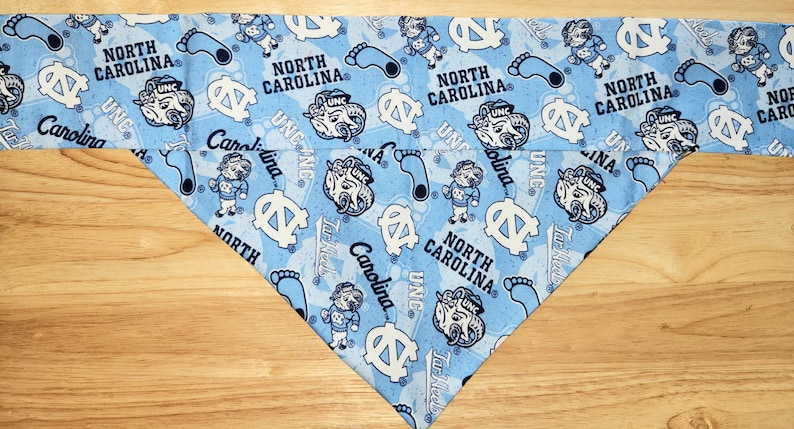 May include: Light blue bandana with a repeating pattern of the words "North Carolina", the letters "UNC", cartoon ram mascots, and footprints. The bandana is folded into a triangle shape.