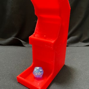 Pode incluir: Uma torre de dados vermelha brilhante em forma de mini-arcade. Um dado transparente de várias faces com números brancos repousa na bandeja. A torre tem um design clássico de arcade com um topo inclinado e detalhes do painel de controle.