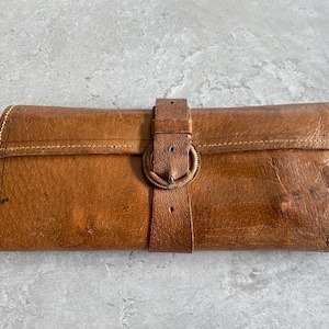 May include: A rectangular tan leather wallet featuring a fold-over flap secured by a vertical leather strap and a circular buckle. The wallet displays visible stitching along the edges and a weathered, textured surface with natural variations in colour.