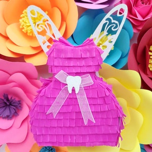 May include: A pink tooth fairy piñata with white wings and a white ribbon with a tooth charm. The piñata is surrounded by colorful paper flowers.