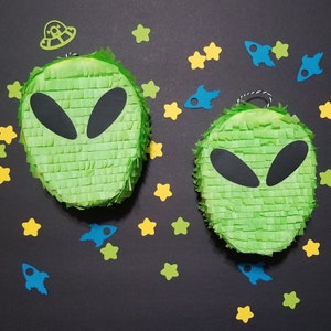 May include: Two green alien-shaped pinatas with black eyes and mouths, surrounded by blue and yellow paper rockets and stars on a black background.