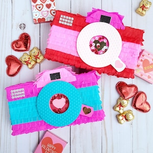 May include: Two camera-shaped piñatas in red, pink, and blue, decorated with fringed paper. The lenses are white and blue with heart-shaped details. Valentine's Day themed with heart-shaped candies and small gift boxes scattered around.