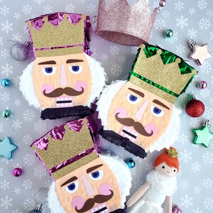 May include: Three Nutcracker pinata decorations with gold crowns and white fur. The pinatas are pink, green, and purple. A pink crown and a white ballerina doll are also in the image.