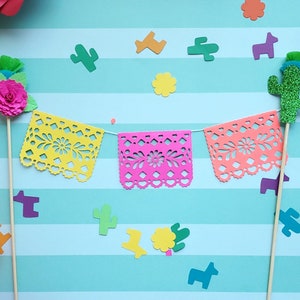 May include: A set of three colorful paper banners with a scalloped edge and intricate cutouts. The banners are strung together with a white string. There are also two paper cutouts of cacti and a flower, and a paper cutout of a donkey. The background is a light blue and white striped pattern.
