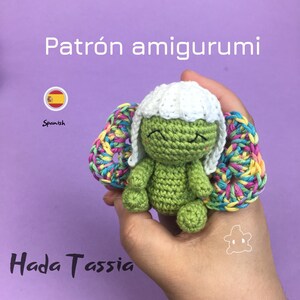 Puede incluir: Una muñeca de hada amigurumi de crochet verde con cabello blanco y alas de arcoíris. La muñeca está sosteniendo sus manos juntas frente a su pecho. La muñeca está sentada en una mano. El texto "Patrón amigurumi" y "Hada Tassia" es visible en la imagen.