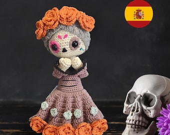 May include: A crocheted doll with a gray skull face, wearing a purple dress with orange flower accents and a crown of orange flowers. The doll is standing on a dark wooden surface next to a white skull and purple flowers.