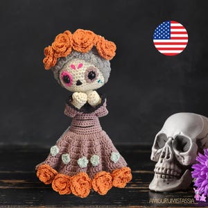 Puede incluir: Muñeca de crochet del Día de Muertos con un vestido morado con detalles de flores naranjas y una cara de calavera gris. La muñeca tiene las manos juntas en posición de oración. La muñeca está sentada sobre una superficie de madera junto a una calavera blanca.