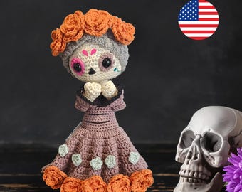 May include: Crocheted Day of the Dead doll wearing a purple dress with orange flower accents and a gray skull face. The doll is holding its hands together in a praying position. The doll is sitting on a wooden surface next to a white skull.