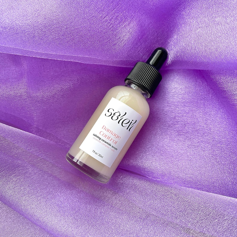 May include: A clear glass bottle with a black dropper top and a white label. The label says "sōlei" in a stylized font and "Damage Control calming ceramide serum  repairs, strengthens, calms" in a smaller font. The bottle is sitting on a purple fabric background.
