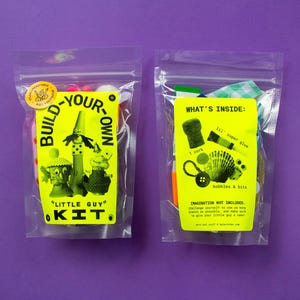 Puede incluir: Dos bolsas de pl&aacute;stico transparente con etiquetas amarillas. Una dice "Build-Your-Own Little Guy Kit" y la otra "What's Inside: bobbles & bits". Las bolsas contienen materiales de manualidades, sobre un fondo morado.