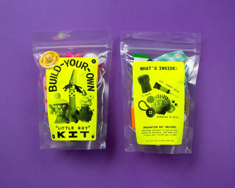 Puede incluir: Dos bolsas de pl&aacute;stico transparente con etiquetas amarillas. Una dice "Build-Your-Own Little Guy Kit" y la otra "What's Inside: bobbles & bits". Las bolsas contienen materiales de manualidades, sobre un fondo morado.