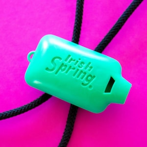 May include: A bright green whistle with the text "Irish Spring" printed on it. The whistle is attached to a black cord and set against a vibrant pink background.