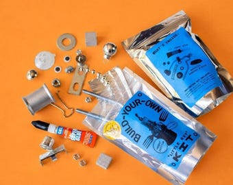 Puede incluir: Un kit de construcción de robot con dos bolsas de aluminio plateado y etiquetas azules. Una etiqueta dice BUILD-YOUR-OWN LITTLE GUY KIT. La otra dice WHAT'S INSIDE. Los componentes sueltos incluyen una bobina de metal, pegamento, tuercas, pernos, arandelas y pequeñas esferas metálicas.