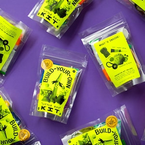 Puede incluir: Varias bolsas de pl&aacute;stico transparente llenas de materiales de manualidades est&aacute;n dispuestas sobre una superficie morada. Cada bolsa tiene una etiqueta amarilla brillante con las palabras "Little Guy Kit" y "Build-Your-Own". Las etiquetas tambi&eacute;n incluyen ilustraciones y texto.