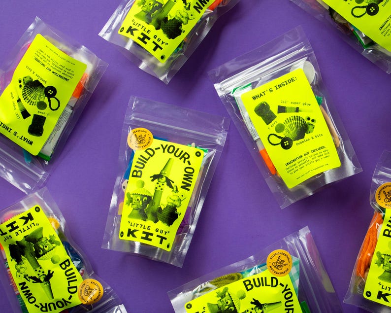 Puede incluir: Varias bolsas de pl&aacute;stico transparente llenas de materiales de manualidades est&aacute;n dispuestas sobre una superficie morada. Cada bolsa tiene una etiqueta amarilla brillante con las palabras "Little Guy Kit" y "Build-Your-Own". Las etiquetas tambi&eacute;n incluyen ilustraciones y texto.