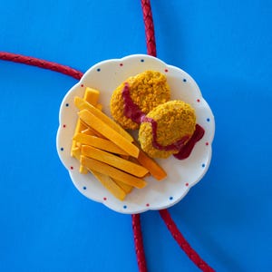 May include: A plate of food with two golden-brown patties drizzled with red sauce and a side of fries. The plate is white with a scalloped edge and red and blue dots. The background is bright blue.