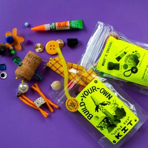 May include: A collection of craft supplies and a DIY kit on a purple background. The kit includes a bottle of glue, a cork, buttons, googly eyes, and other small items. The kit's label reads "Build Your Own Little Guy Kit."