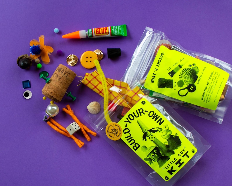 Puede incluir: Un conjunto de materiales de manualidades y un kit de bricolaje sobre un fondo morado. El kit incluye un tubo de pegamento, un corcho, botones, ojos saltones y otros objetos peque&ntilde;os. La etiqueta del kit dice "Build Your Own Little Guy Kit."