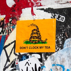 May include: A yellow sticker with a black and white image of a coiled snake with the text "Don't Clock My Tea" and a smaller text "Don't Tread On Me" underneath the snake.