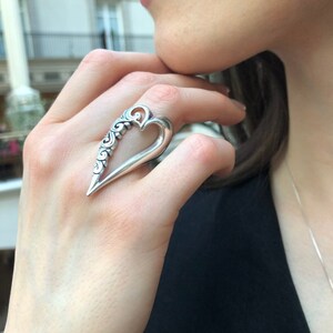 May include: A silver ring with a heart-shaped design. The heart is open and has intricate details on the outside.