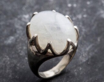 May include: A silver ring featuring a large, oval, milky white gemstone. The ring's setting has a crown-like design, with pointed prongs holding the stone. The ring is set against a dark, textured background.