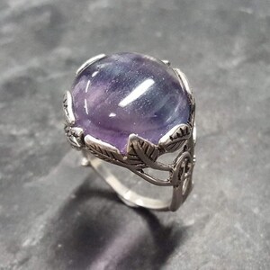 May include: A silver ring with a large, oval-shaped purple and blue fluorite gemstone set in a leafy design.