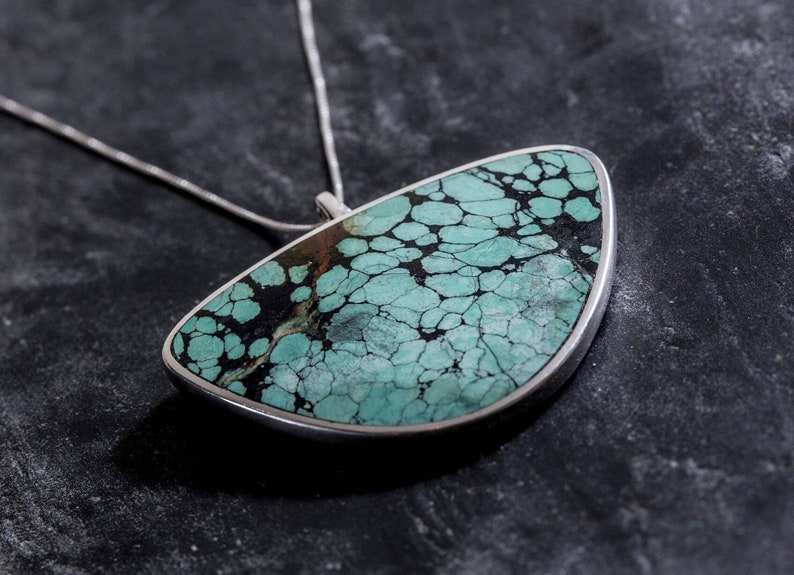 May include: A silver necklace with a large, turquoise pendant. The pendant is a teardrop shape and has a mottled, green and black pattern.