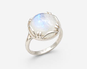 May include: A silver ring featuring a large, round, iridescent moonstone. The stone displays a soft, milky white color with blue and purple flashes. The ring's setting has a decorative, openwork design.