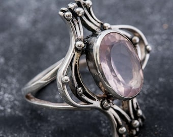 May include: A silver ring featuring a light pink oval gemstone. The ring has an elaborate design with curved lines and small, spherical details. The gemstone is set in a bezel, and the ring is against a dark backdrop.