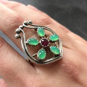 May include: A silver ring with a green gemstone flower design. The ring has a red gemstone in the center of the flower.