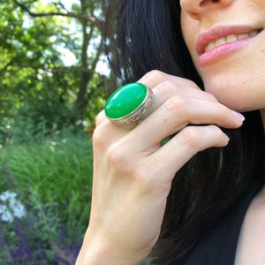 Könnte beinhalten: Ein großer, ovaler Ring mit einem grünen Edelstein, der in Silber gefasst ist. Der Ring hat komplizierte Details um den Stein herum.