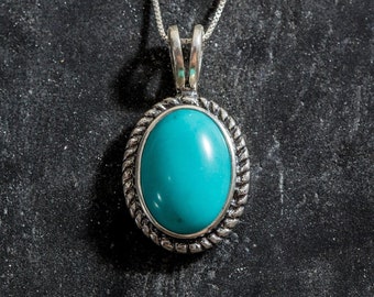 Puede incluir: Collar con colgante de turquesa ovalado con un borde plateado estilo cuerda y una cadena delicada. La piedra azul verdosa brillante está engastada en un engaste de metal plateado. El collar se muestra sobre una superficie oscura y texturizada.