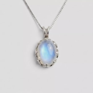 Könnte beinhalten: Eine silberne Halskette mit einem ovalen, schillernden Mondsteinanhänger. Der Anhänger ist in einer dekorativen, gewellten Lünette gefasst. Die Kette ist eine filigrane, quadratische Gliederkette. Der Mondstein zeigt eine blau-weiße Farbe.